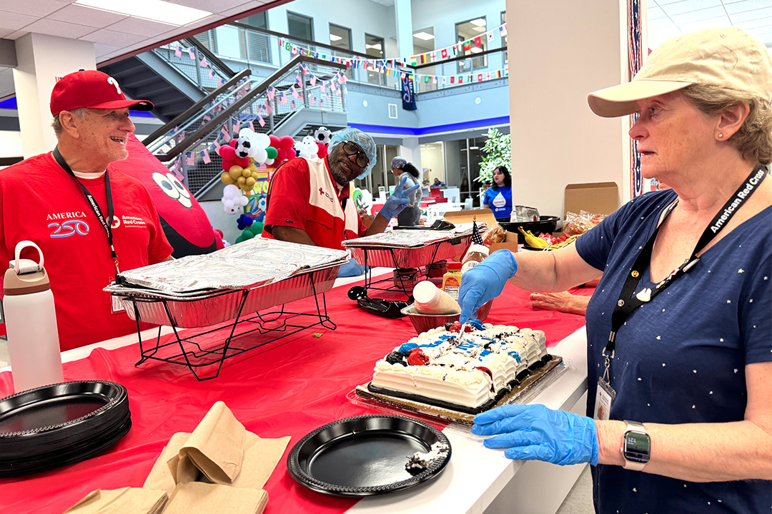 Three smiling mass care feeding team volunteers stand behind a long table covered in a red tablecloth indoors. On the right, Karen Westermann wears a blue Red Cross polo shirt, a khaki visor, and blue medical gloves as she carefully slices a decorated sheet cake. To her left, Mike Kucenic wears a red Red Cross "America 250" t-shirt and a red baseball cap, while Kevin Savage stands smiling in the background. Packages of black paper plates, napkins, and other food service items are laid out on the table.