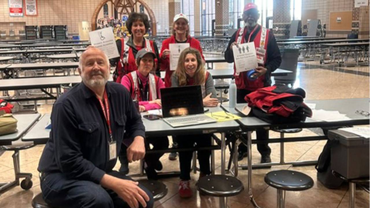 Five American Red Cross disaster responders pose around a registration table inside a large school cafeteria being used as an emergency shelter. Three volunteers wearing Red Cross vests stand behind the table holding accessibility and shelter information signs, while two others sit at the table with a laptop and supplies. Rows of cafeteria tables and benches fill the background.