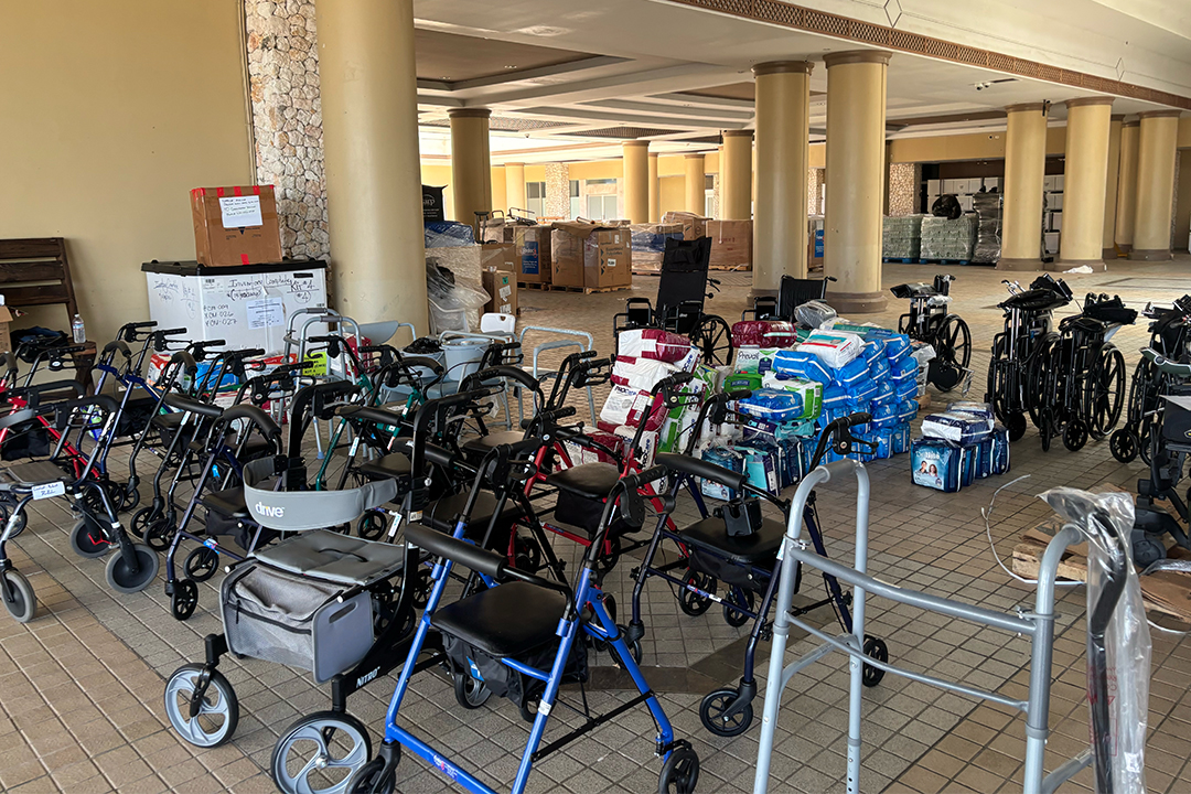 A wide-angle view inside a large, open room with concrete pillars, densely filled with rows of black wheelchairs and metal walkers. Stacks of cardboard boxes and bottled water are visible in the background.