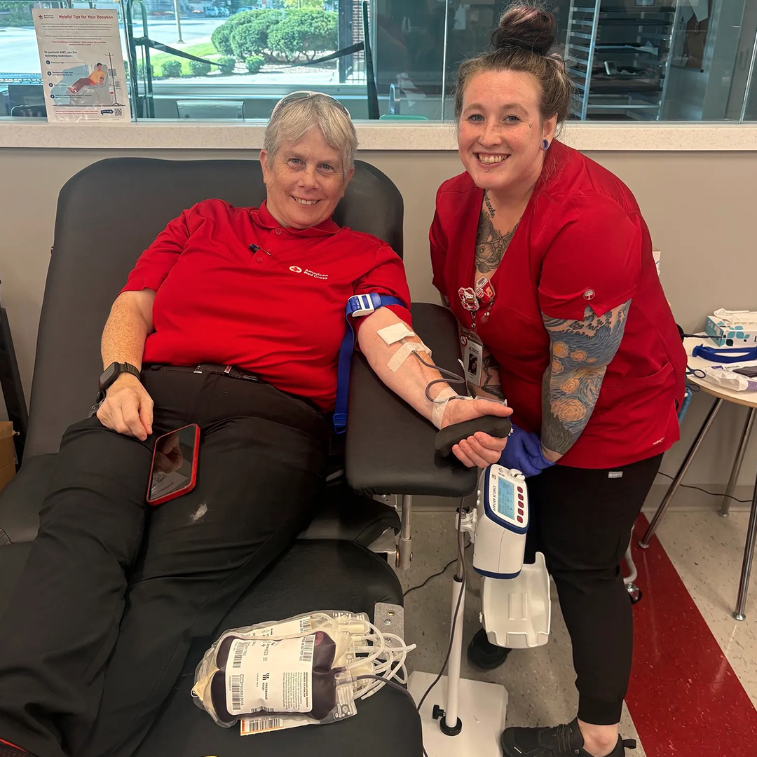 Red Cross volunteer Heidi Dampman sitting in a donor chair smiling next to a phlebotomist while donating blood.