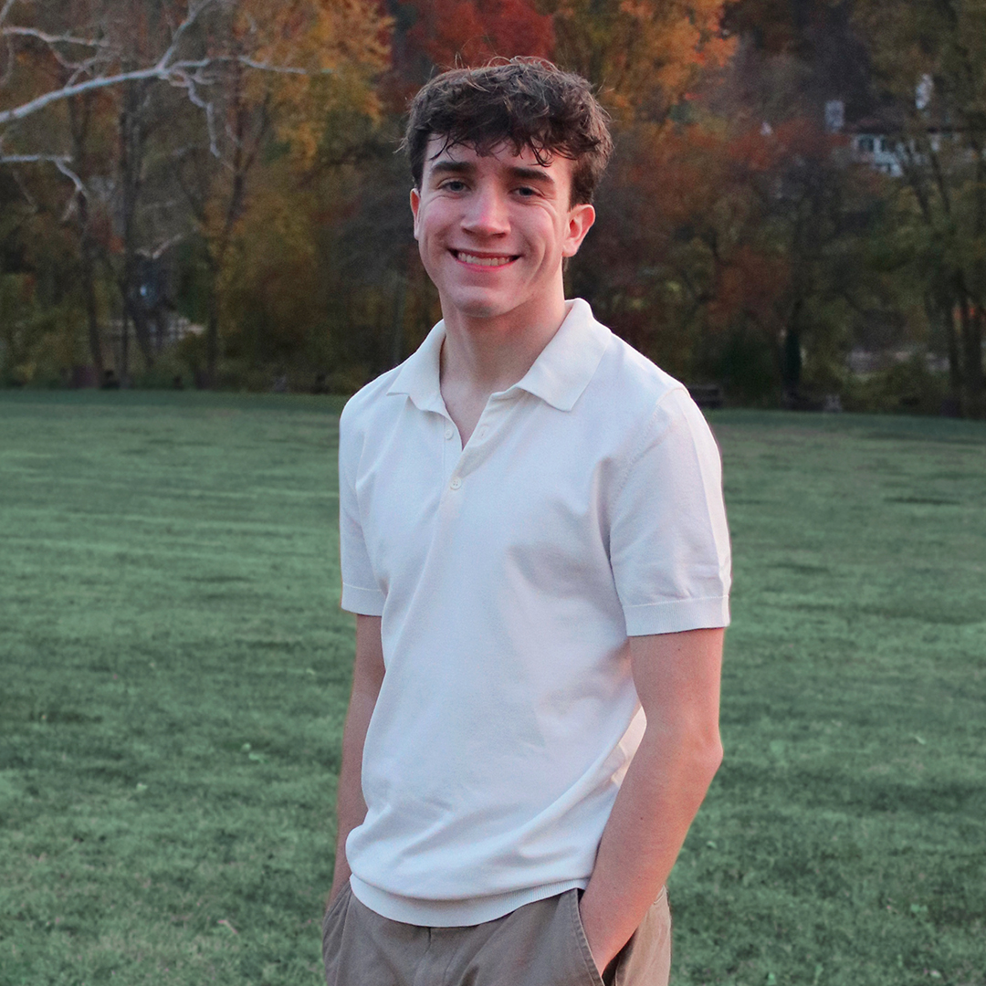 James Frank smiling outdoors in a grassy park, wearing a white polo shirt and beige pants with hands in his pockets.