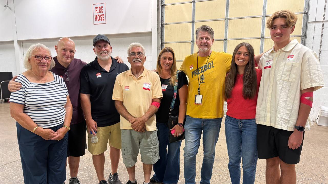 A group of eight people pose for a photo indoors, including blood donors and radio co-hosts Steve Morrison and Preston Elliot, standing side-by-side in a row and smiling at the camera.