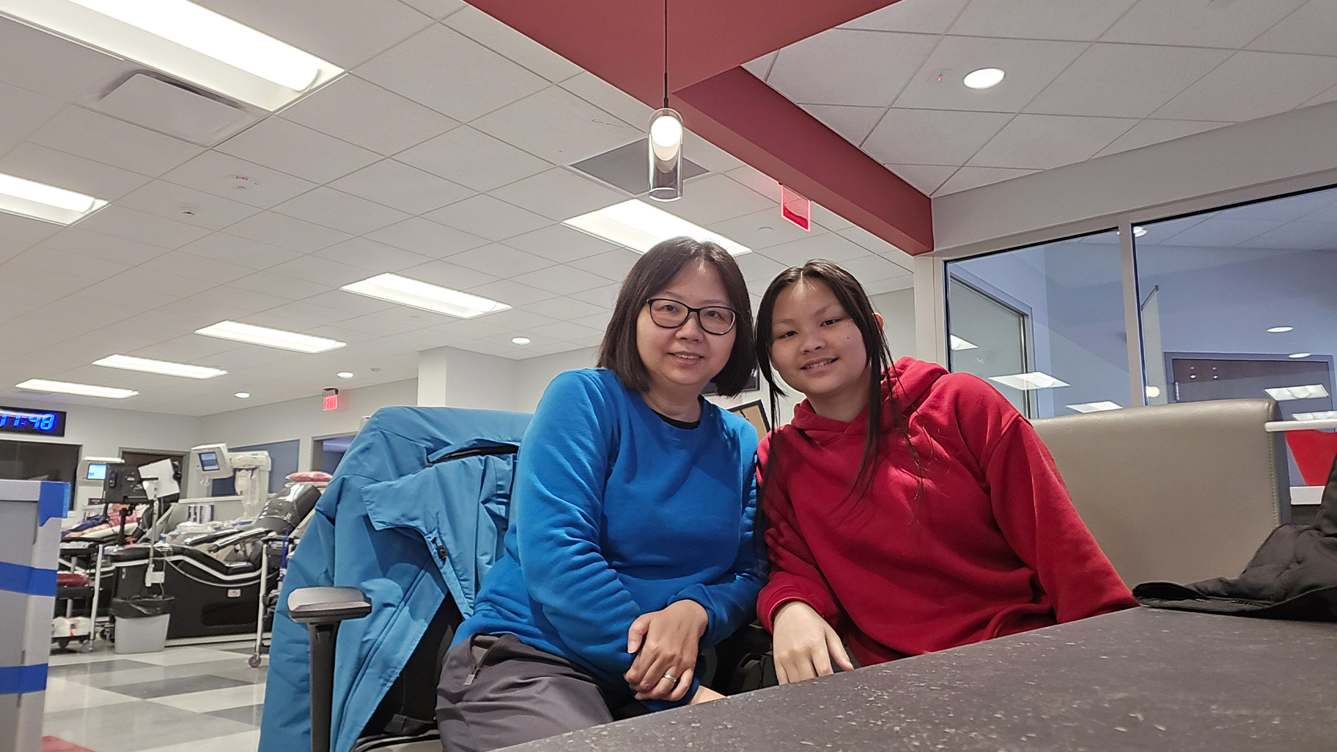 Mother and daughter Red Cross volunteers Jane and Juniper sit at a table with blood donation chairs visible behind them. 