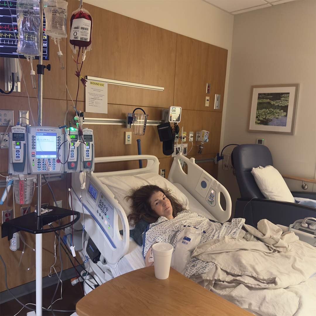 Photo of a woman in a hospital bed with a blood bag and IVs hanging next to her as she receives a blood transfusion.