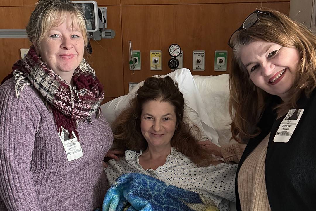 Two caucasian women stand next to a caucasian women laying in a hospital bed covered with a blue blanket.