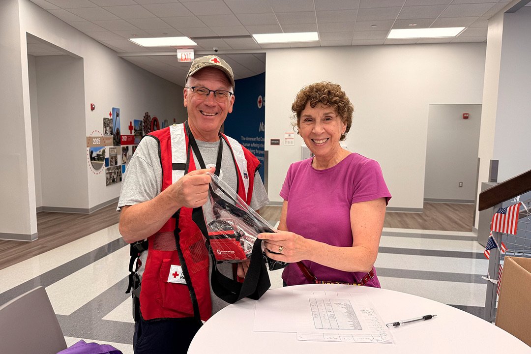 Indoors at a white round table, Ambassador Terri Whiteford, wearing a purple shirt, smiles as she hands a clear plastic swag bag filled with items to shuttle driver John Timalonis. John stands smiling on the left, wearing a red American Red Cross volunteer vest over a white t-shirt, a baseball cap, and glasses. A sign-in sheet and a pen sit on the table between them.