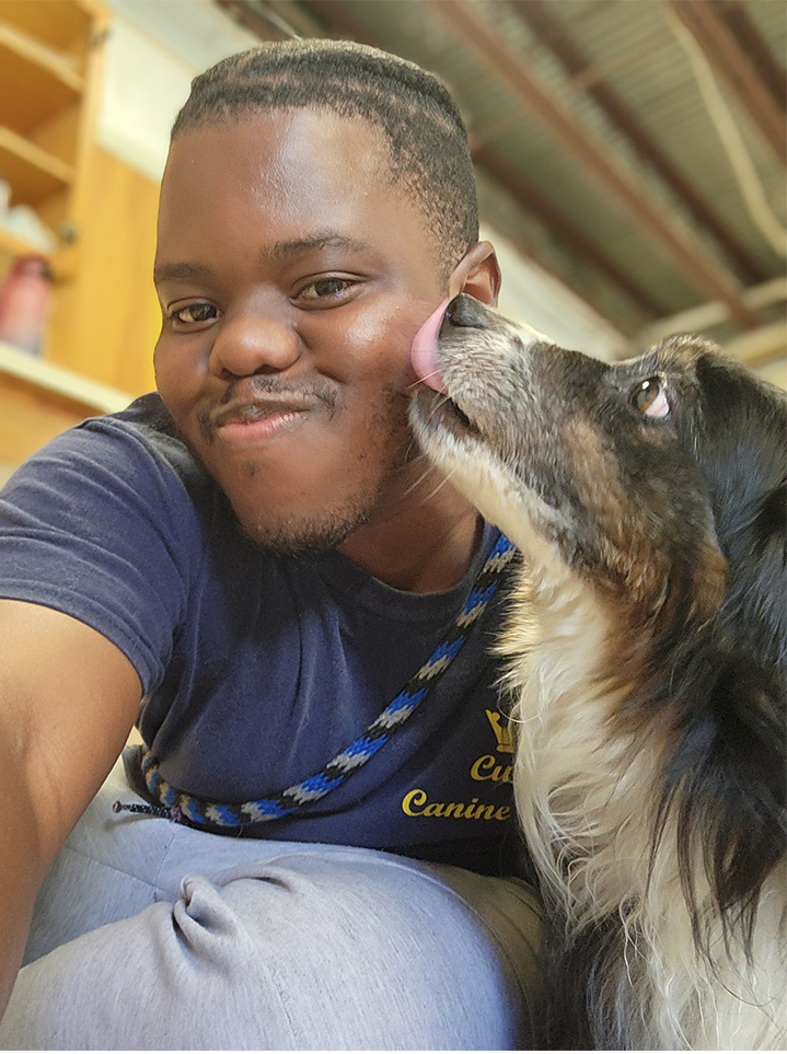 Photo of a young Black man bending down while a brown and white dog licks his face.