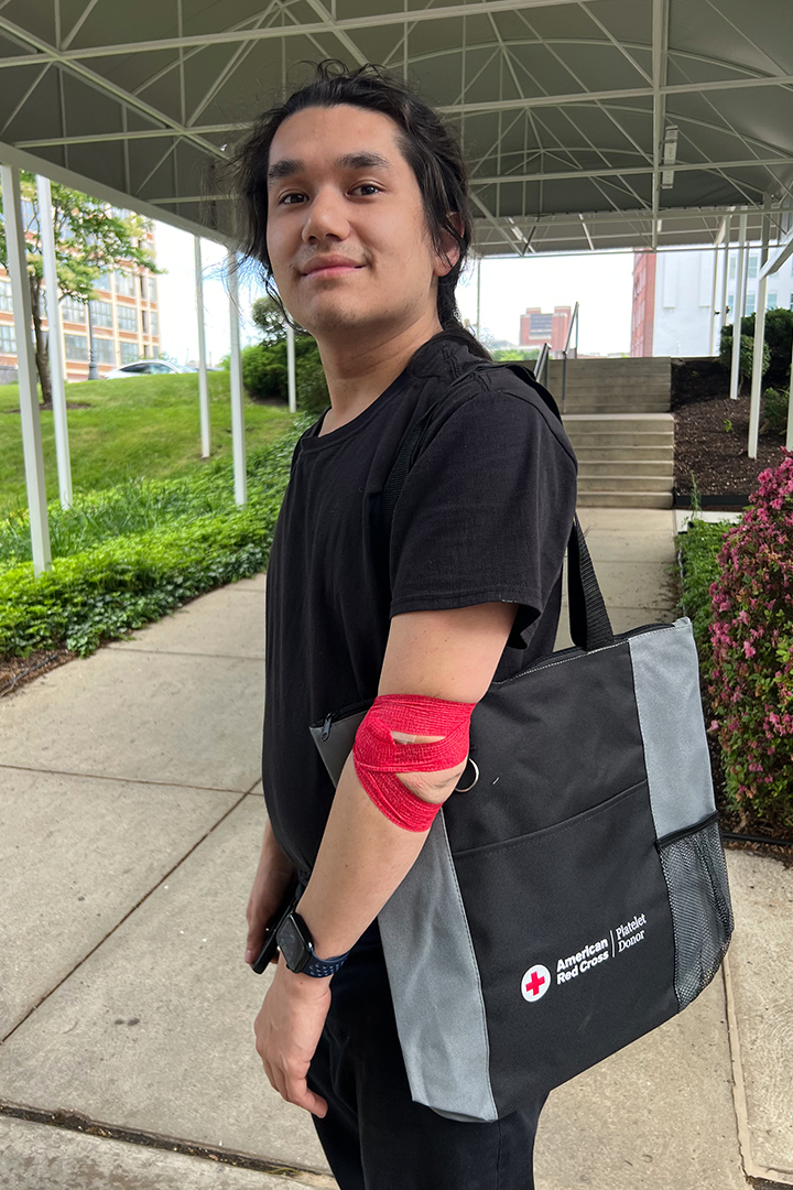 Kai Kwong stands outside the Red Cross Spring Garden Campus in Philadelphia after donating platelets. He has tape on his arm and his holding a tote bag that says American Red Cross Platelet Donor.