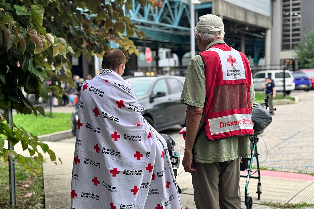 Red Cross volunteer Richard Strayves stands with a woman wrapped in a Red Cross blanket on the sidewalk in front of a parking lot.