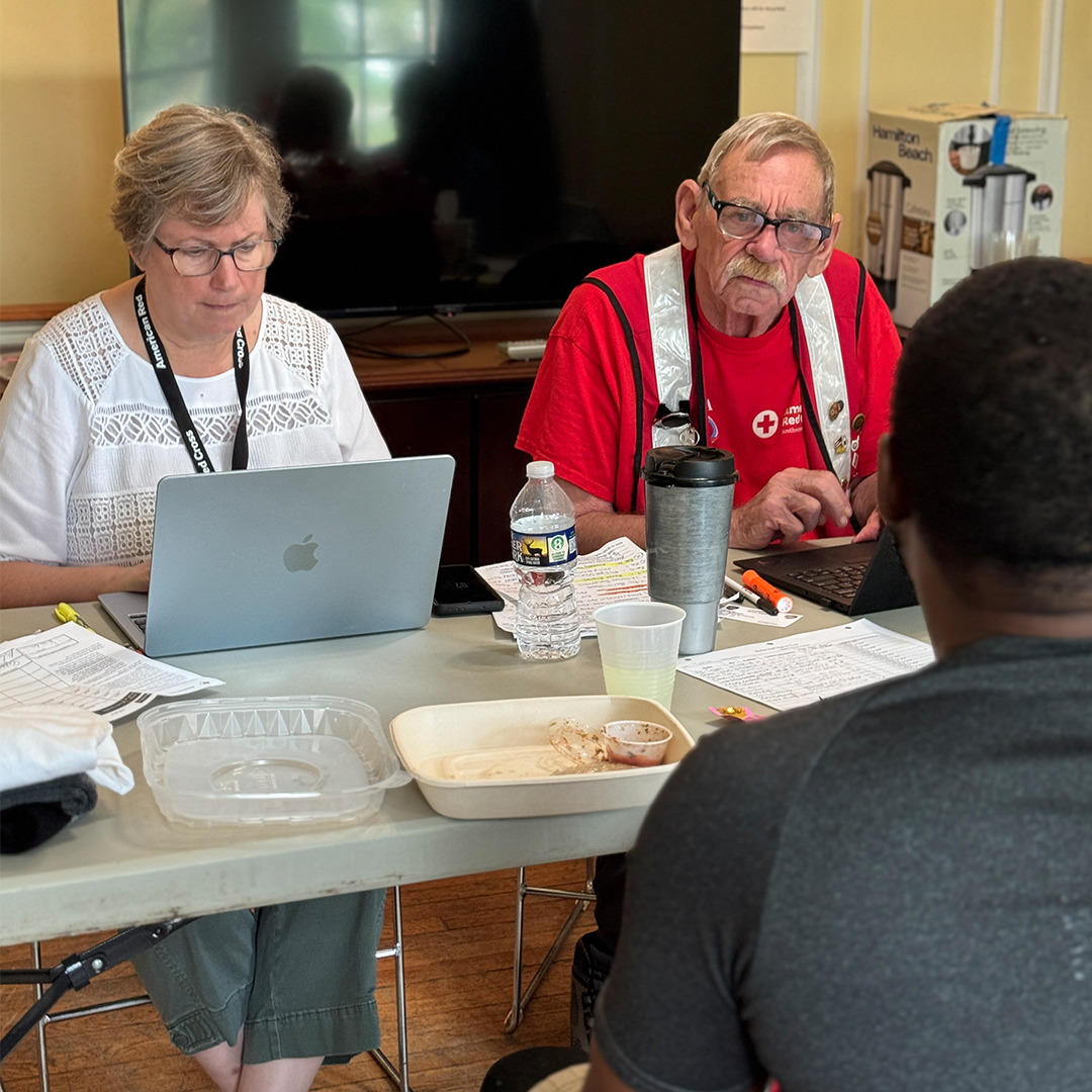 Two Red Cross volunteers sitting at a table with laptops, assisting a person seated with their back to the camera in a shelter setting.