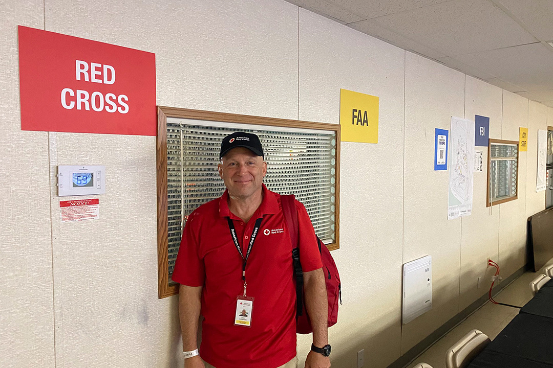 Disaster Action Team Responder LeRoy Whitehead stands smiling inside a long, narrow mobile emergency operations center (EOC) trailer. He wears a red American Red Cross polo shirt, a black baseball cap, khaki pants, and a volunteer badge around his neck. A bright red "RED CROSS" sign is mounted on the wall to his left, and further down the hallway, yellow and blue signs mark "FAA" and "FBI" stations above long rows of desks and equipment.