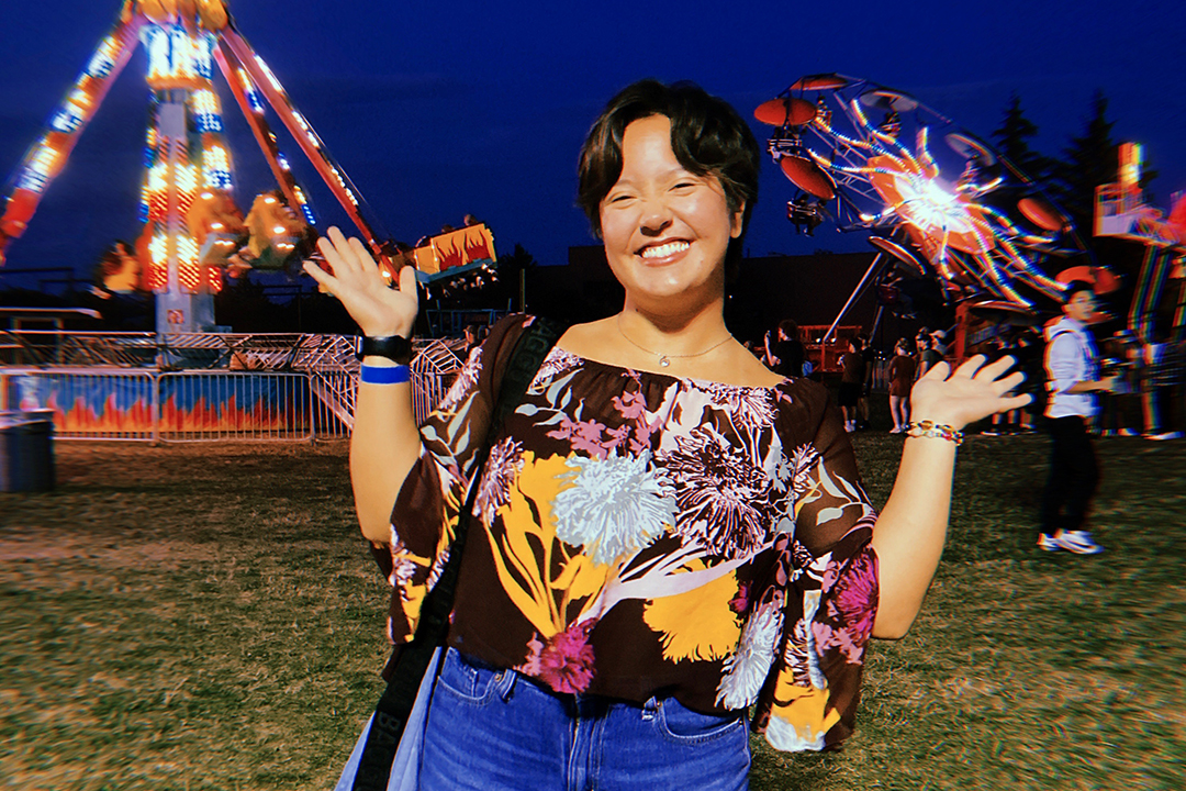 A teenage girl stands in front of a carnival ride with her hands in the air.