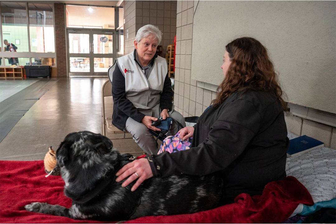 Photo of Linda Fahey talking with a woman sitting on a shelter cot with a large dog. 