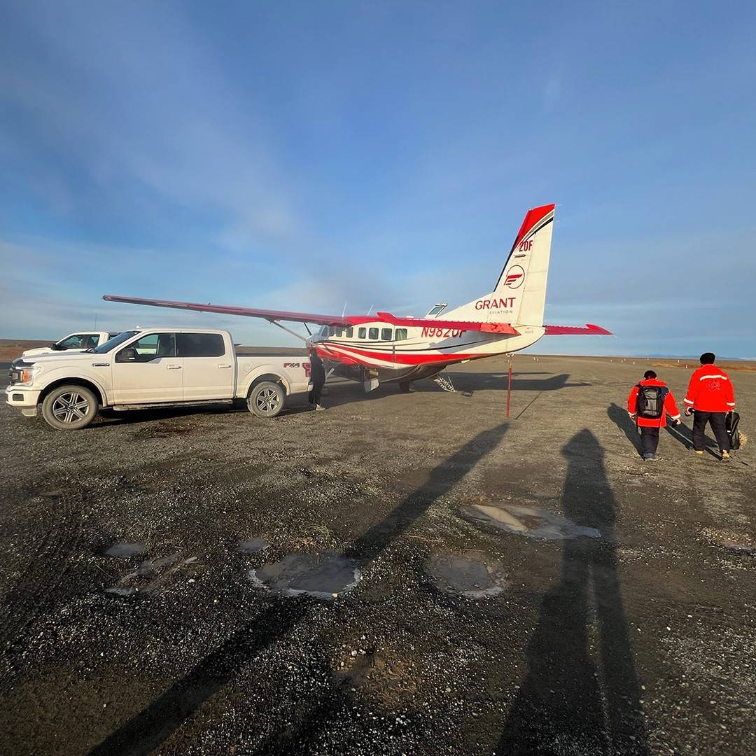 A photo of a small red and white airplane sitting behind a white truck in the Alaskan tundra as two people wearing Red Cross vests walk toward it,.