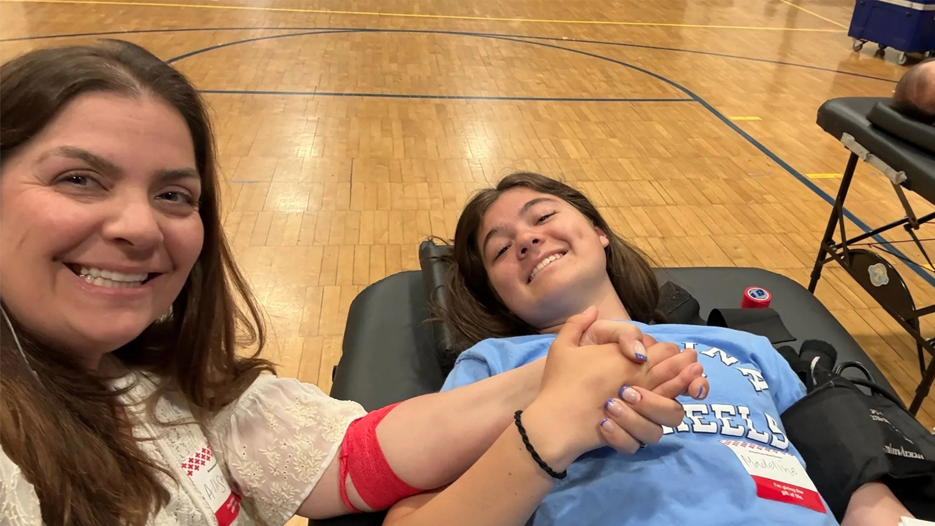 Maddie Mafodda lies on a cot in a high school gymnasium, smiling and holding her mother Allison's hand while donating blood during a Red Cross blood drive.