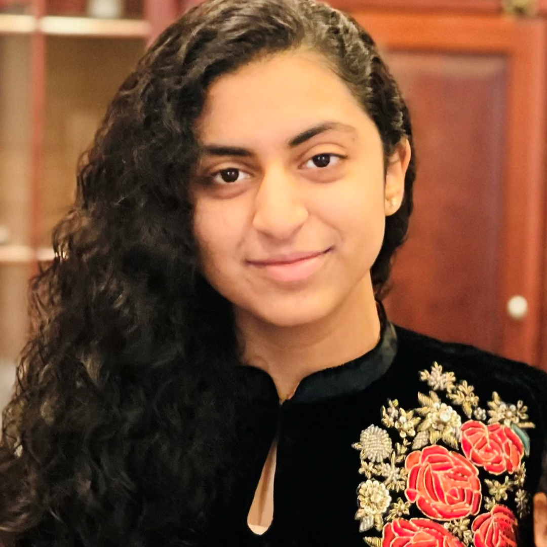 Malaika Virk smiling in a black top with intricate red and orange floral embroidery, with long dark curly hair styled to one side.