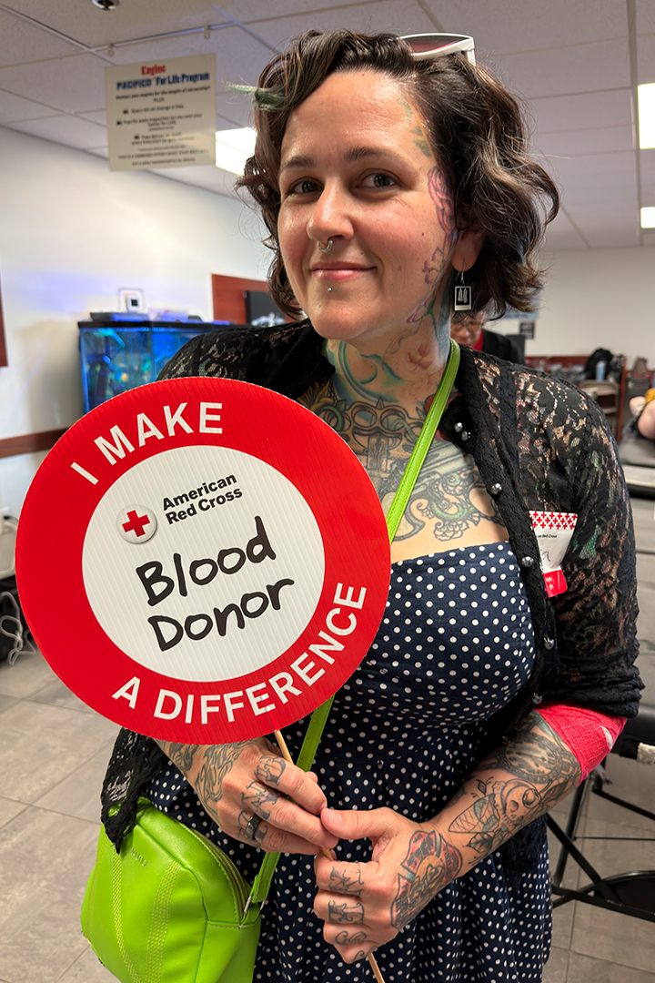 A smiling woman with short, wavy dark hair and tattoos on her arms and chest stands indoors, holding a large circular red sign that reads "I MAKE A DIFFERENCE - American Red Cross Blood Donor". She is wearing a dark, polka-dot top with a lace cardigan over it.