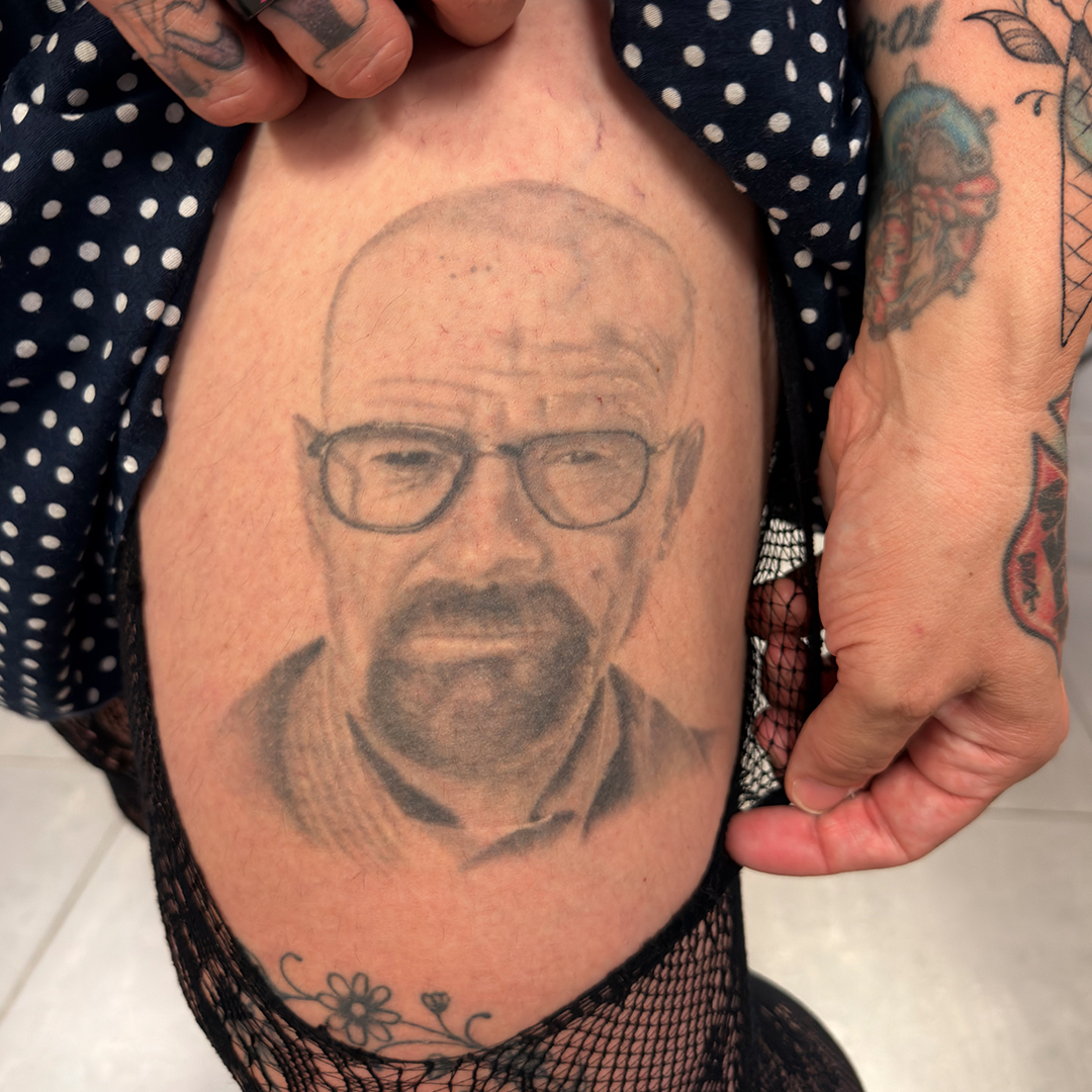 A close-up black-and-grey portrait tattoo of the fictional character Walter White from "Breaking Bad" on a person's thigh. The character is depicted with his signature shaved head, goatee, and wire-rimmed glasses. The person is holding up the hem of a dark polka-dot garment to reveal the tattoo, showcasing other colorful tattoos on their forearms and hands.