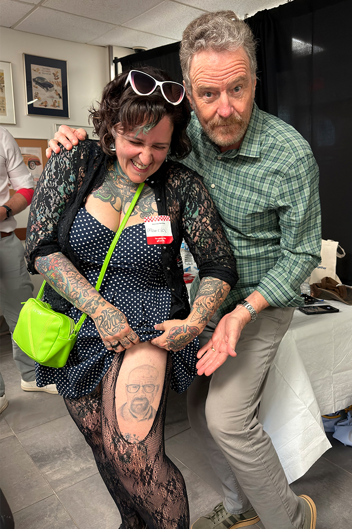 A smiling woman with short, wavy dark hair and extensive arm tattoos lifts the hem of her polka-dot dress to show actor Bryan Cranston a tattoo of his "Breaking Bad" character, Walter White, on her thigh. Cranston, wearing a green plaid shirt, leans in next to her with a humorous expression, gesturing toward the tattoo with his hand.