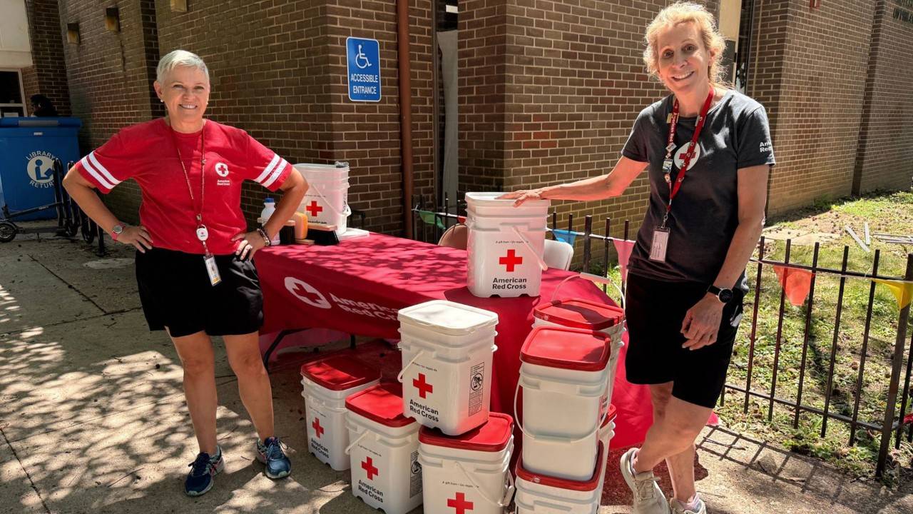 American Red Cross volunteers Barbara McGarvey and Dinae Norris smiling while standing next to stacked Red Cross cleanup kits outside the Wynnfield Library.