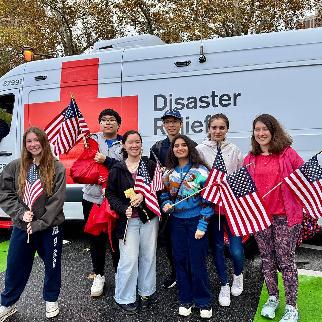 A group of Lower Merion High School Red Cross Club students smiling and holding small American flags in front of a white American Red Cross Disaster Relief vehicle.