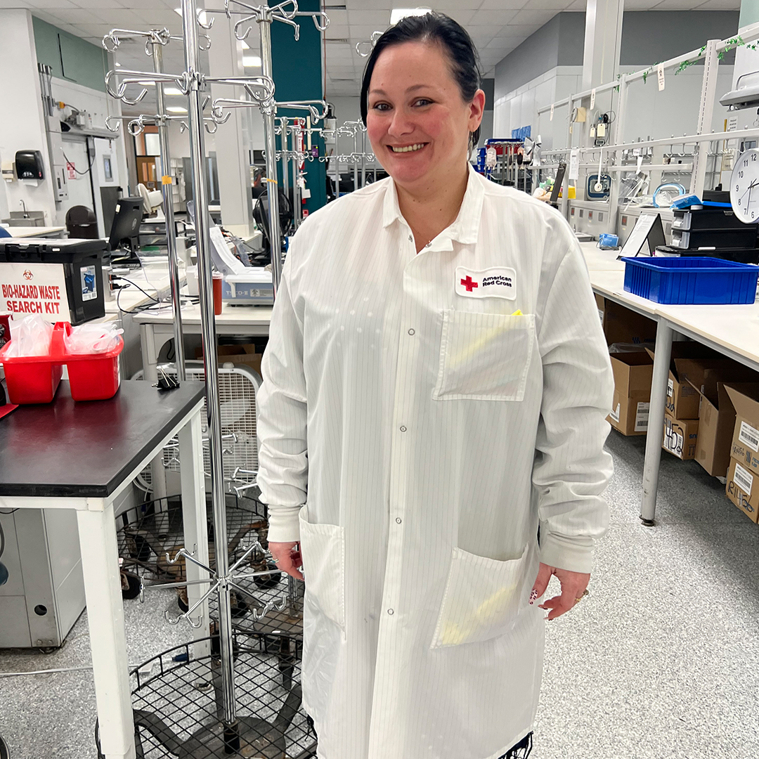 Lead Laboratory Technician Nora Magras smiles while wearing a white lab coat and blue patterned shirt inside the American Red Cross Manufacturing, Quarantine & Labeling Lab in Philadelphia, surrounded by lab equipment and supplies.