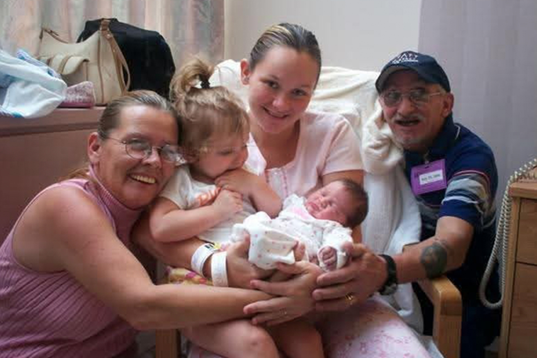 Nora Magras is shown in a hospital room smiling alongside her mother Gerry Redding and late father Michael Redding while holding her newborn baby Hailey, as two-year-old Felisha looks on.
