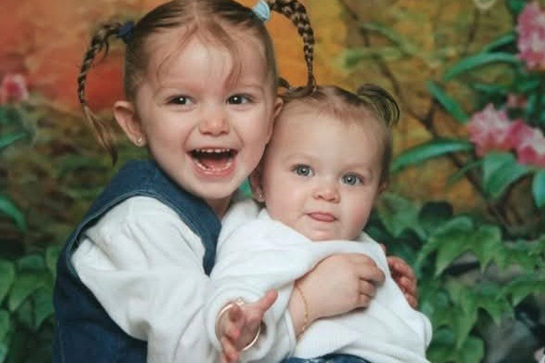 Two-year-old Felisha has her arms wrapped around her baby sister Hailey, who is sitting on her lap, while both girls smile for the camera.