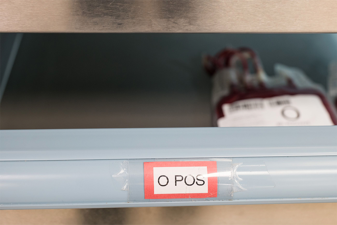 A close-up shot inside a hospital refrigerator shelf showing a plastic bag filled with blood. A prominent red and white label on the front of the shelf directly below the blood bag reads "O POS".