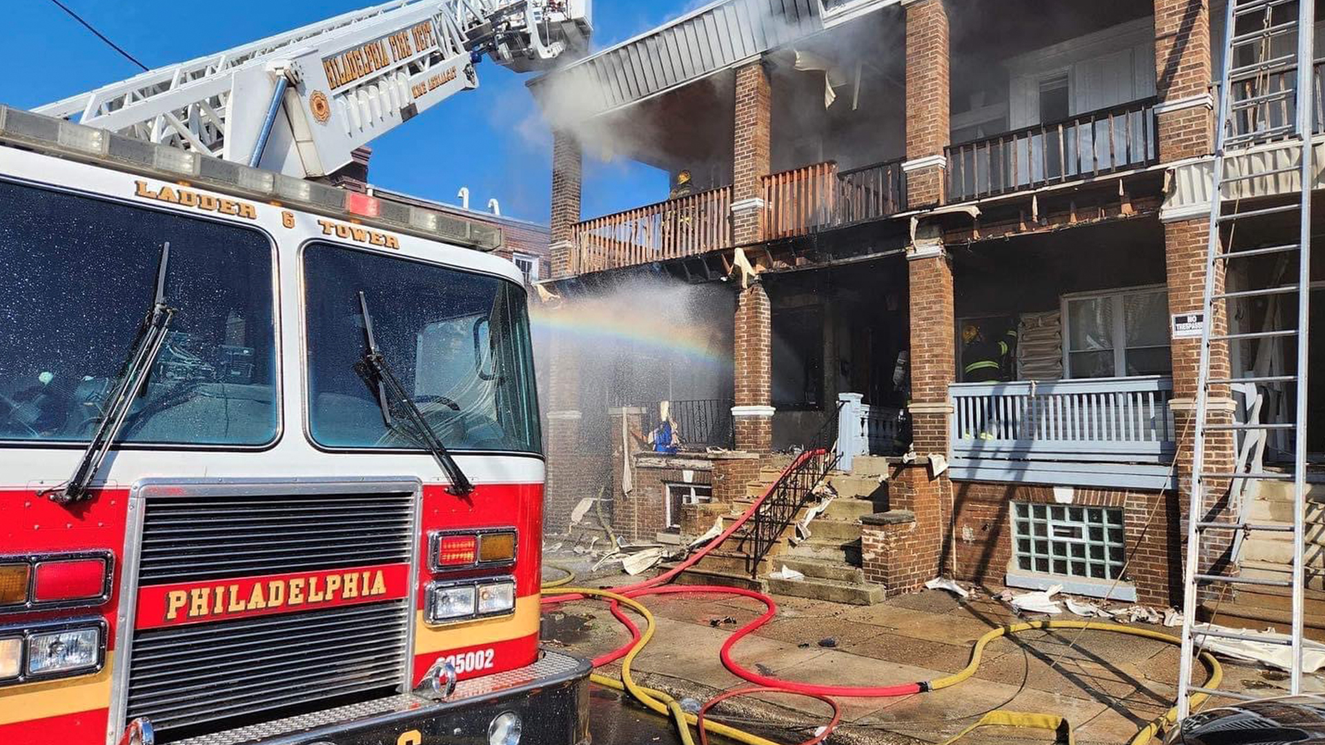 A Philadelphia Fire Department truck is parked in front of a multi-family residential building with fire damage. Orange and yellow hoses are on the ground. 