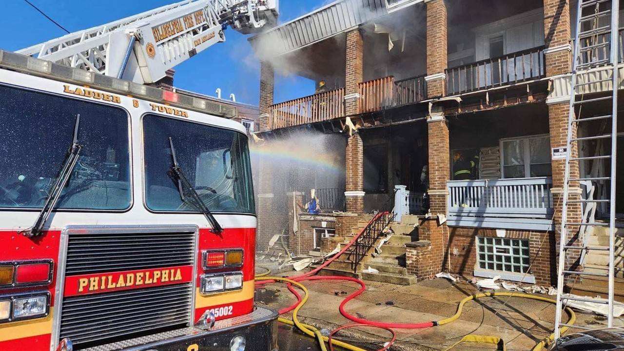 A Philadelphia Fire Department truck is parked in front of a multi-family residential building with fire damage. Orange and yellow hoses are on the ground. 