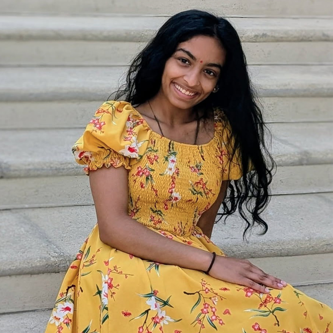 Prapti Patel, a 2026 Downingtown East High School graduate, sitting on stone steps and smiling while wearing a floral yellow midi dress with puffed short sleeves.