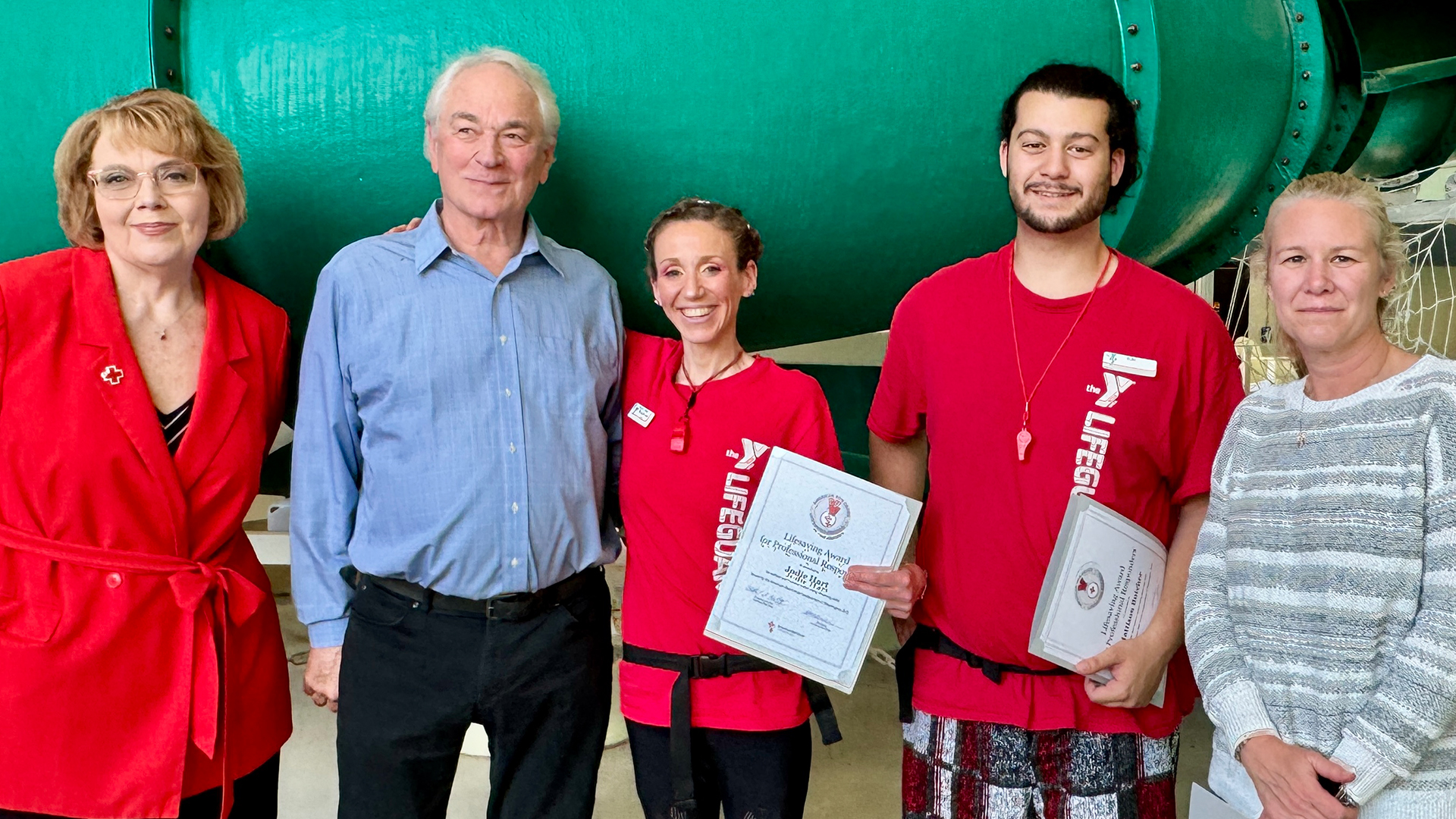 5 people stand in front of a water slide. 2 people are wearing YMCA lifeguard shirts and are holding certificates.