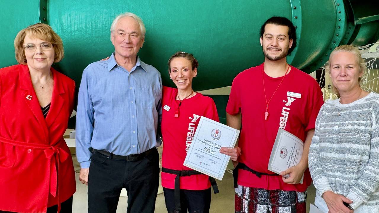 5 people stand in front of a water slide. 2 people are wearing YMCA lifeguard shirts and are holding certificates.
