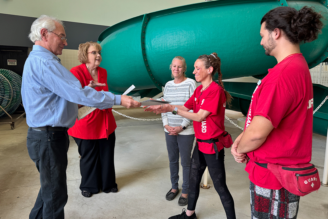 An older man hands a piece of paper to a young woman wearing a lifeguard T-shirt while 3 other people look on. Everyone is standing in front of a water slide.