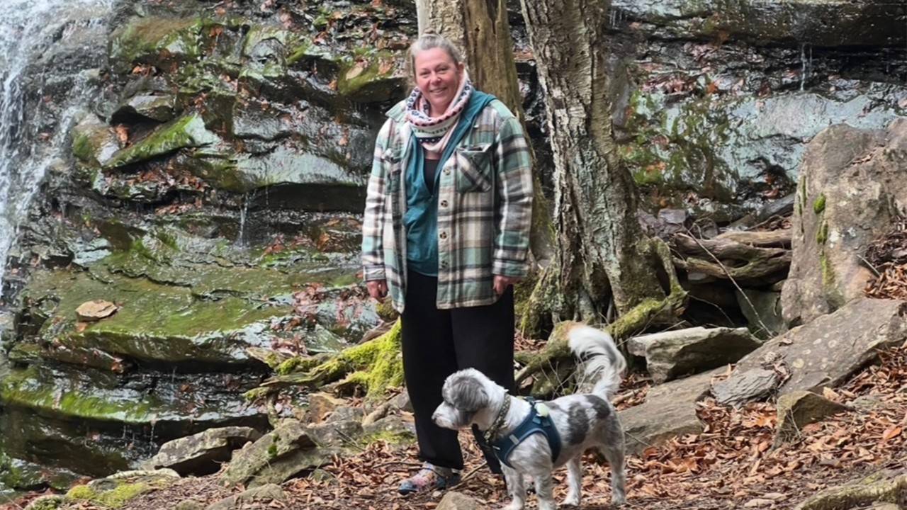A woman walks in the woods with a small gray and white dog.