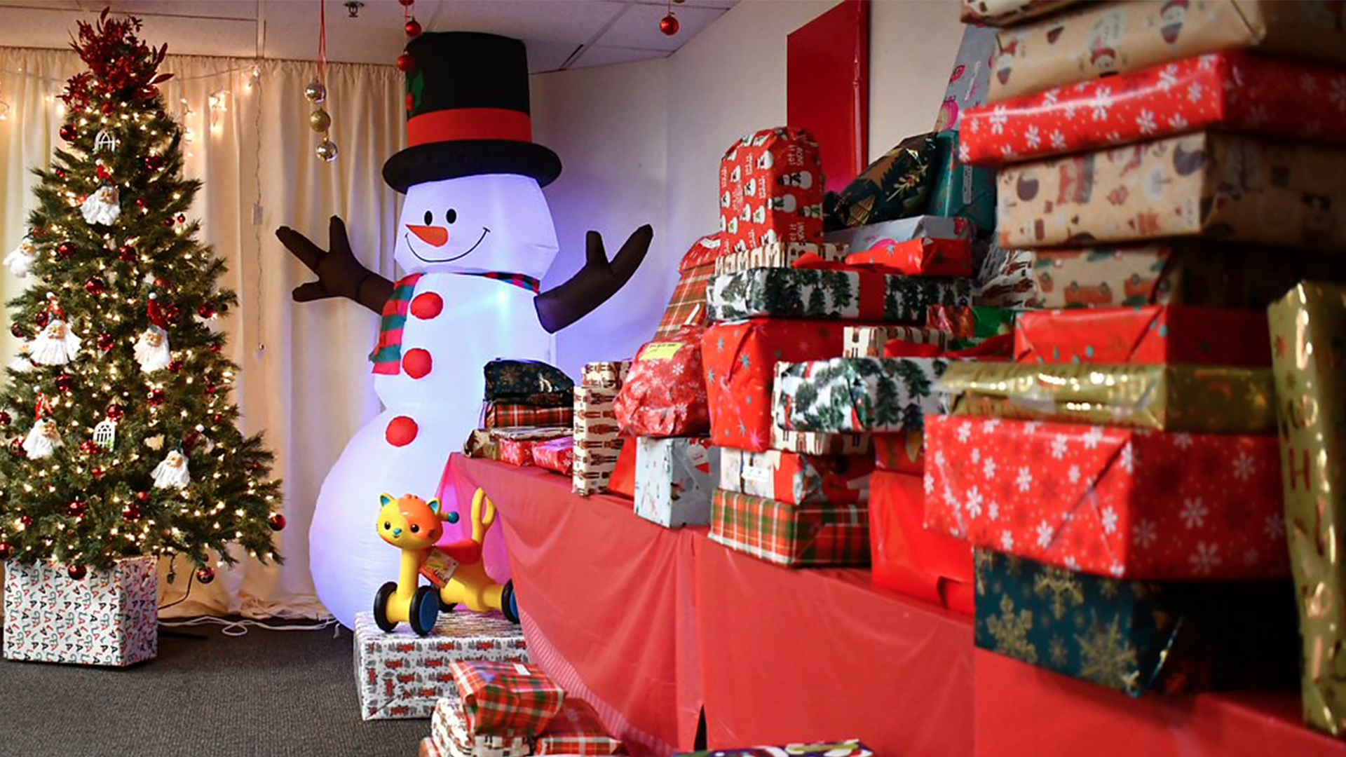 Dozens of wrapped Christmas gifts sit on a long table. A large inflatable snowman and decorated Christmas tree sit at the end. 