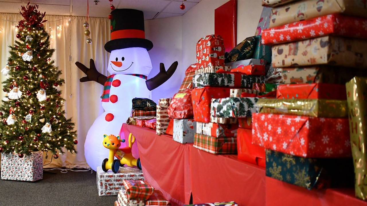Dozens of wrapped Christmas gifts sit on a long table. A large inflatable snowman and decorated Christmas tree sit at the end. 