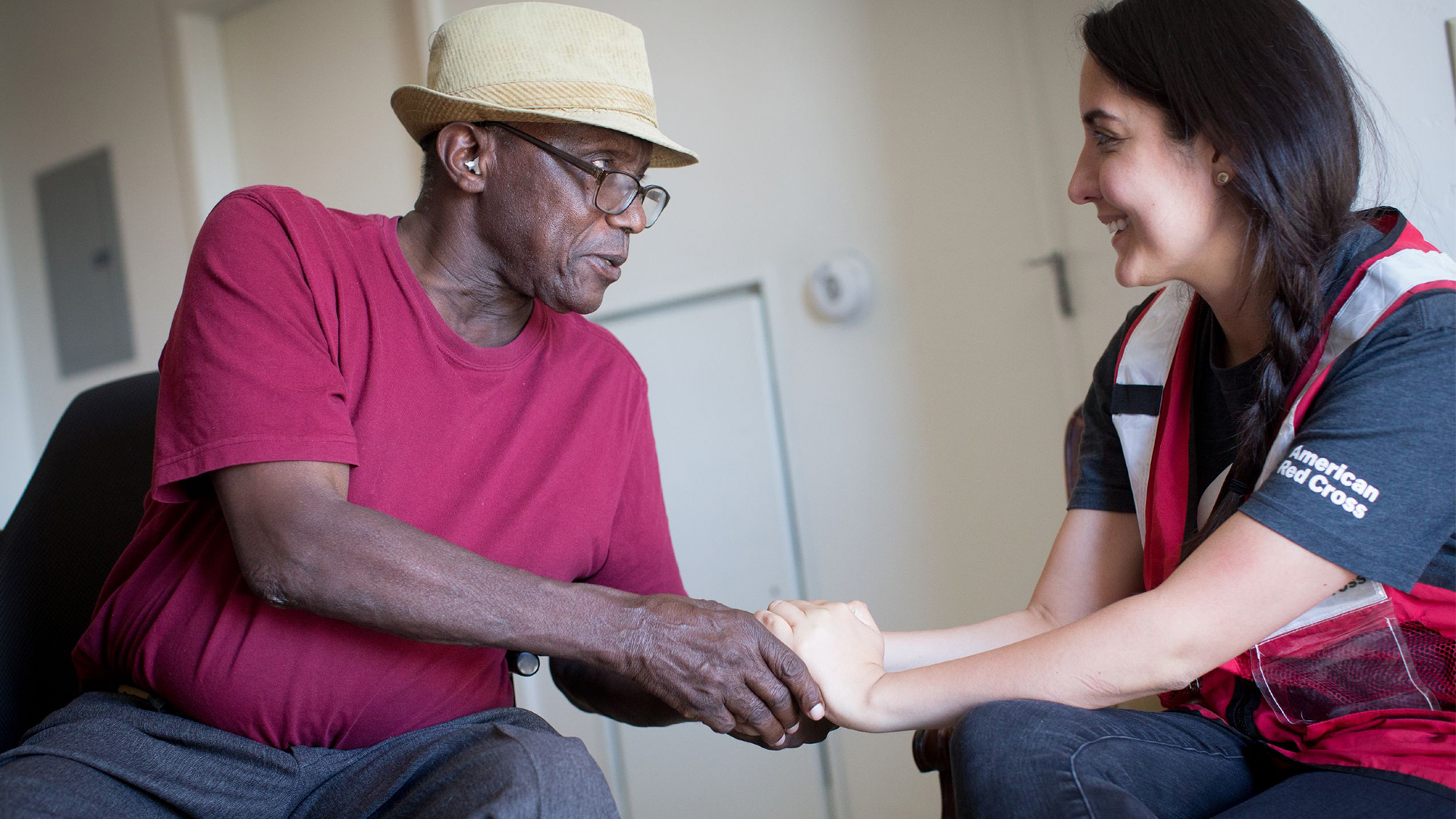 A young woman in a Red Cross vest is pictured grasping an African American man's hand. 