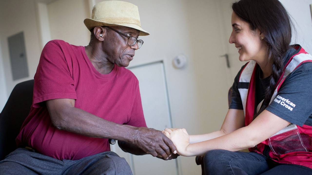 A young woman in a Red Cross vest is pictured grasping an African American man's hand. 