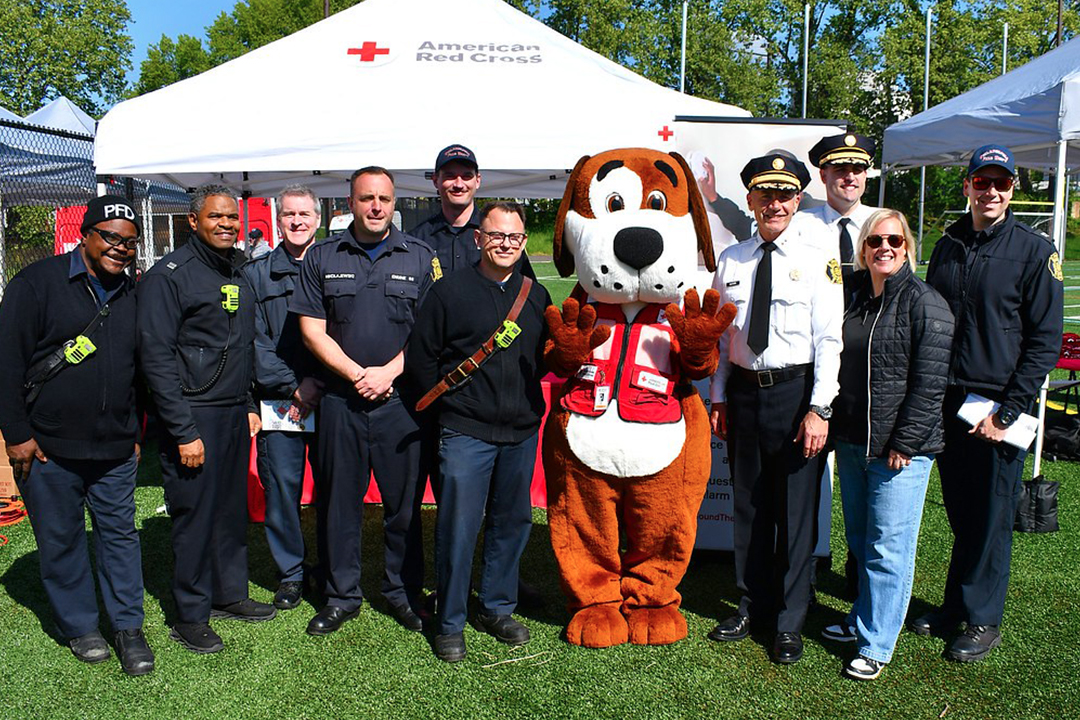 Group photo of members of the Philadelphia Fire Department standing in front of an American Red Cross canopy with Red Cross CEO Jennifer Graham and mascot Fred Cross. who is a lifesize plush dog.