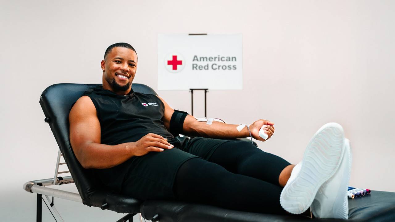 Dressed in black with white sneakers, Saquon Barkley sits on a blood donation table with the American Red Cross logo behind him. 