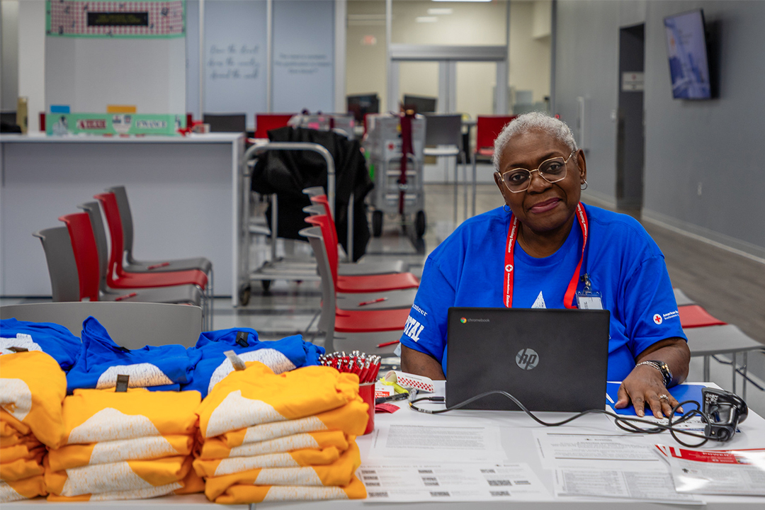 A smiling Red Cross volunteer, Crystal Baytop, sits indoors behind a white registration table wearing a bright blue shirt, glasses, and a lanyard. She is typing on an HP laptop, with stack of yellow and blue "DONATE" T-shirts arranged on the left side of the table in front of her.