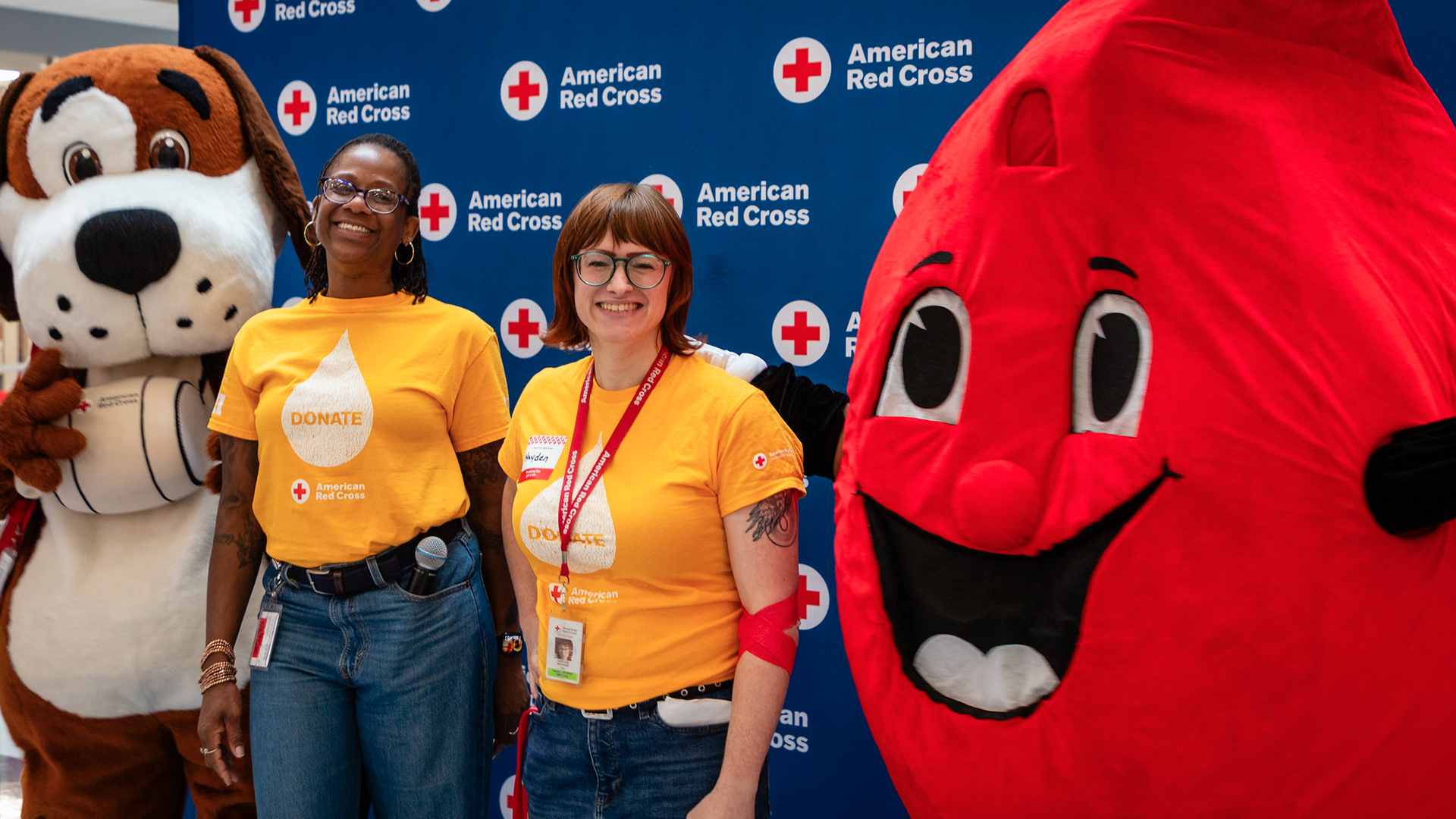 Two Red Cross staff members in yellow "DONATE" shirts pose with a dog mascot and a blood drop mascot at a blood drive.