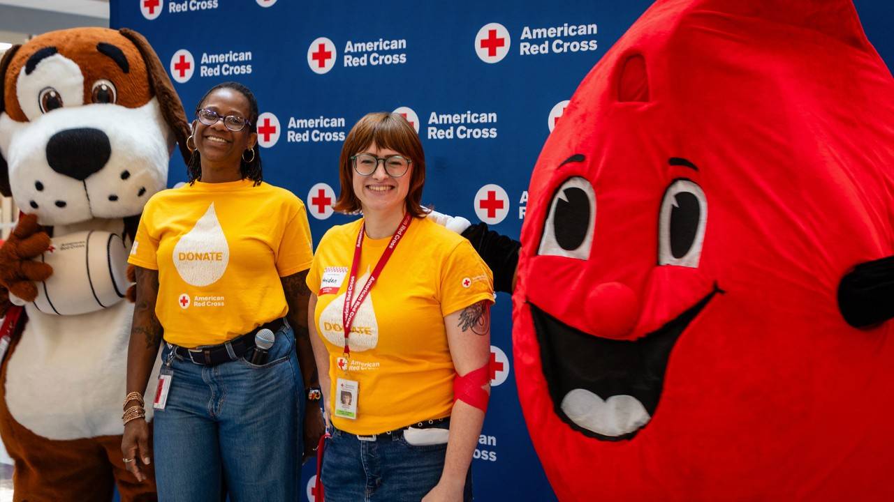 Two Red Cross staff members in yellow "DONATE" shirts pose with a dog mascot and a blood drop mascot at a blood drive.