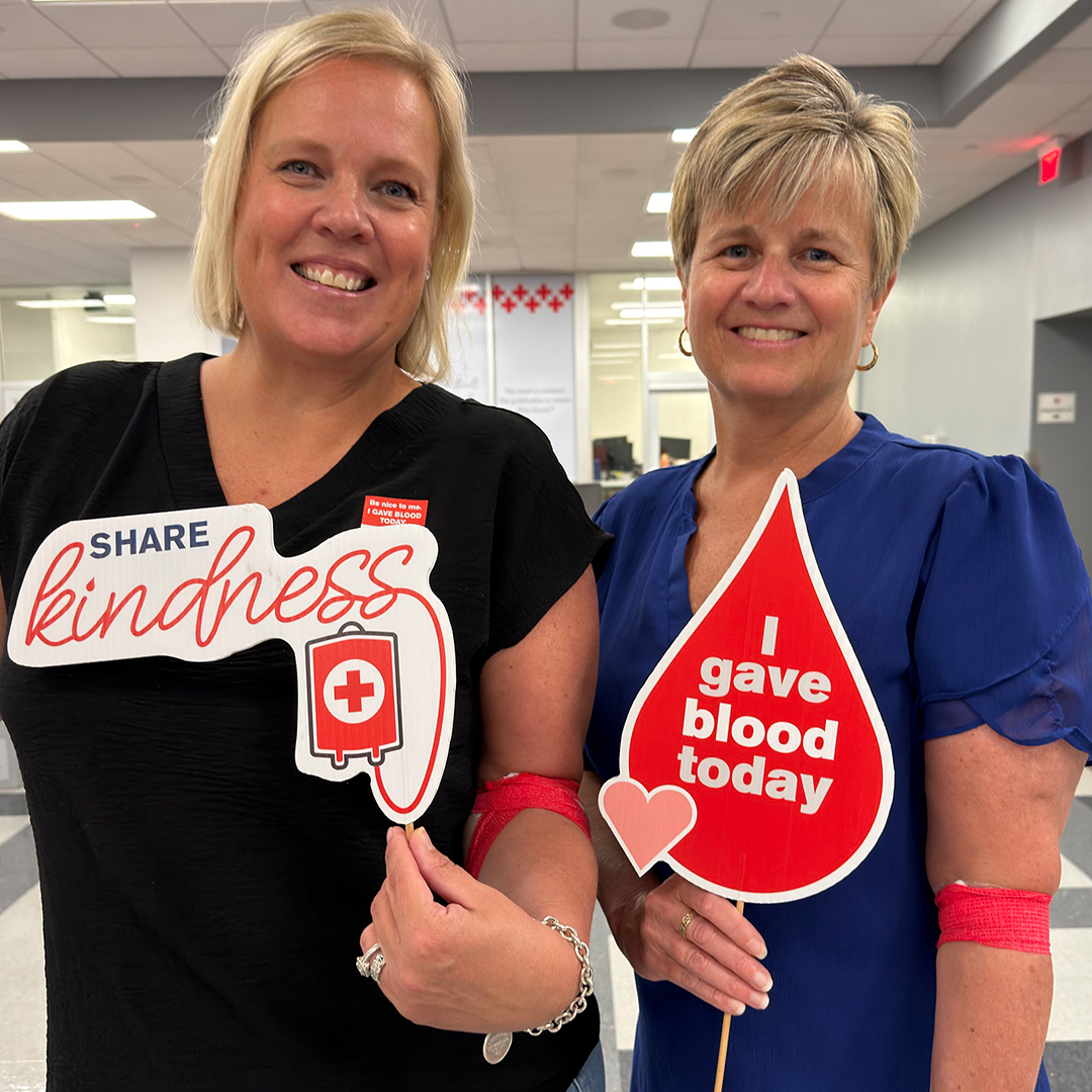 Two smiling women hold Red Cross "Share kindness" and "I gave blood today" props at a blood drive.