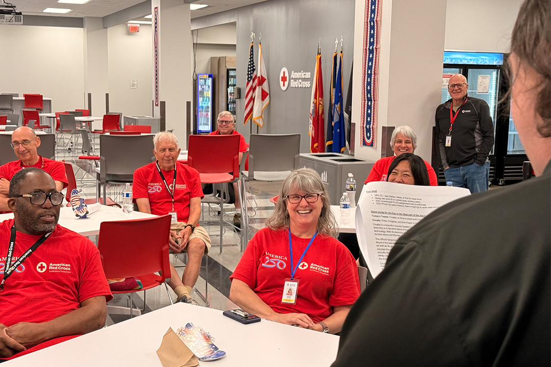 American Red Cross volunteers wearing red shirts are seated and gathered in a bright room for a briefing at the Spring Garden campus, with flags and an American Red Cross logo displayed in the background.