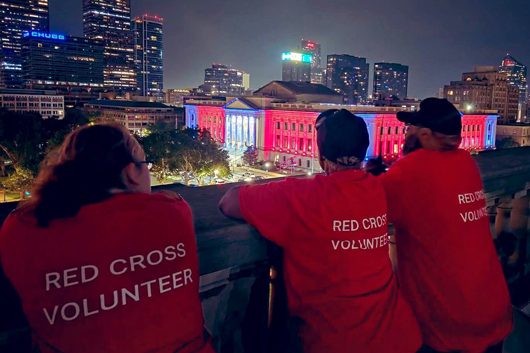Three American Red Cross volunteers wearing red shirts labeled "RED CROSS VOLUNTEER" stand looking out toward a glowing city skyline at night with illuminated colorful buildings in the distance.