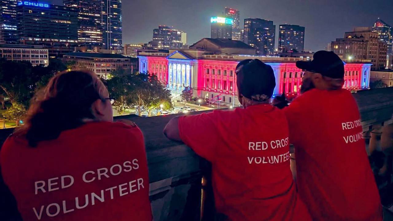 Three American Red Cross volunteers wearing red shirts labeled "RED CROSS VOLUNTEER" stand looking out toward a glowing city skyline at night with illuminated colorful buildings in the distance.