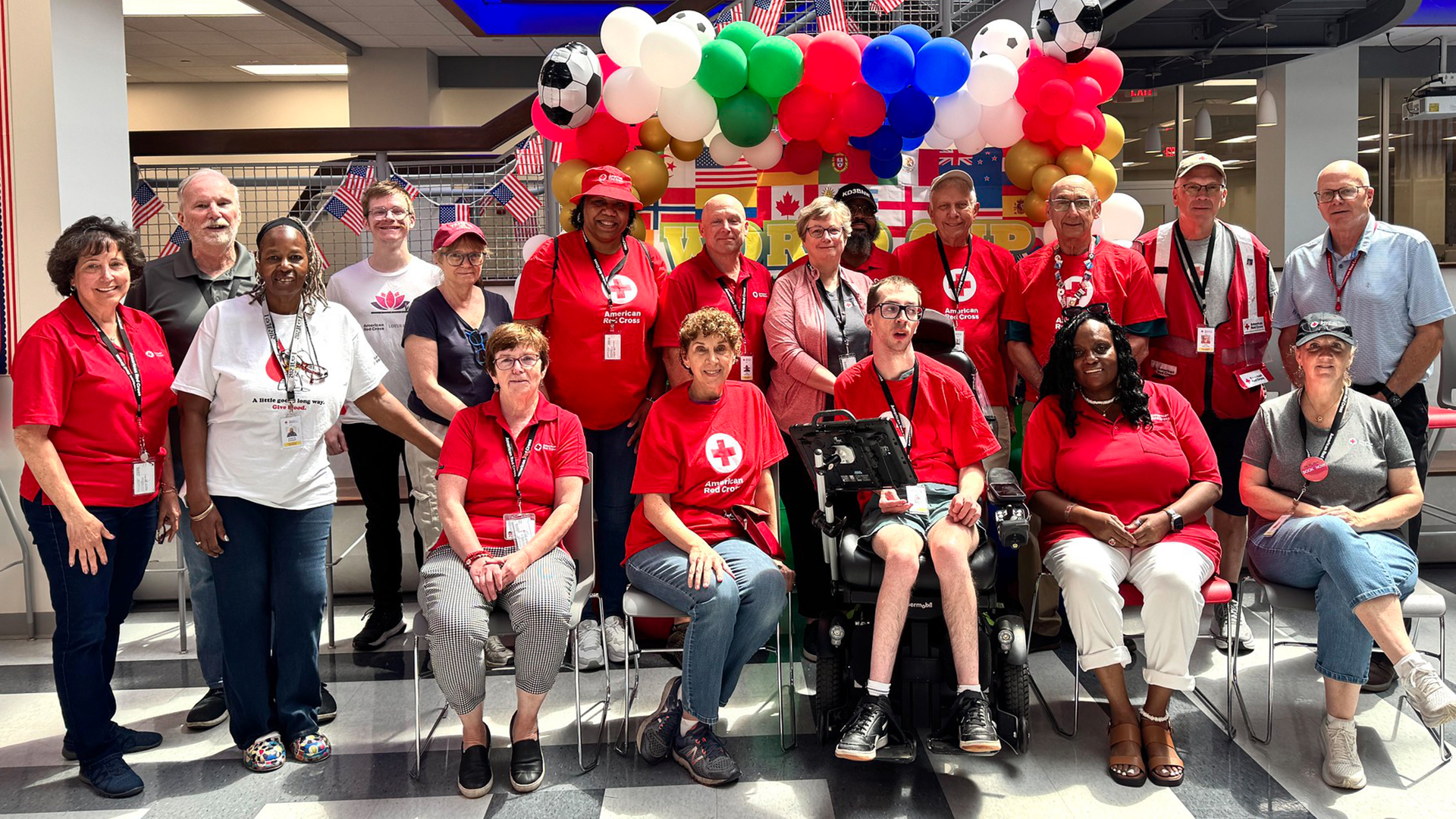 A large group of about fifteen smiling American Red Cross volunteers and staff pose together indoors for a group photo. Most are wearing bright red Red Cross T-shirts, while a few wear polo shirts or casual attire. They are gathered in a brightly lit hallway or atrium decorated with festive red, white, and blue balloon arches and hanging international flags in the background.